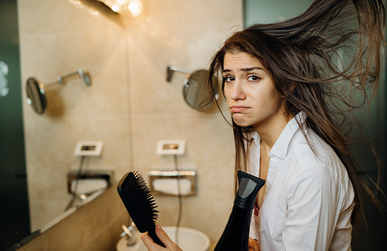 Mujer con cabello graso sosteniendo un secador de pelo y un cepillo y haciendo una cara triste